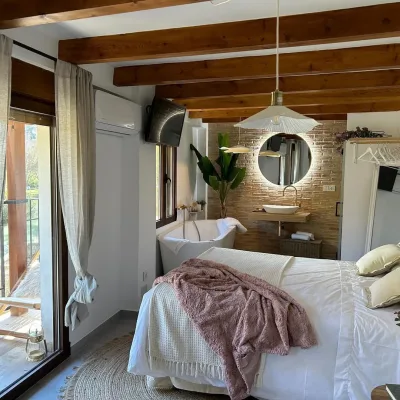 Imagen de la casa Coquette, en la que se ve la amplica cama, la bañera hidromasaje al lado de la cama y el lavamanos al lado, con un espejo con luz redondo encima del lavamanos, también se puede apreciar la televisión y el aire acondicionado.